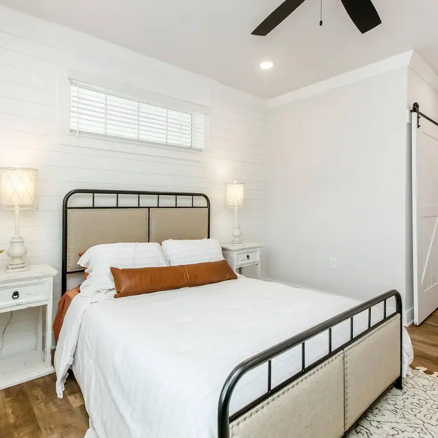 Modern bedroom with a metal bed frame, white bedding, two lamps, and sliding barn door. Warm wood floors complete the decor.