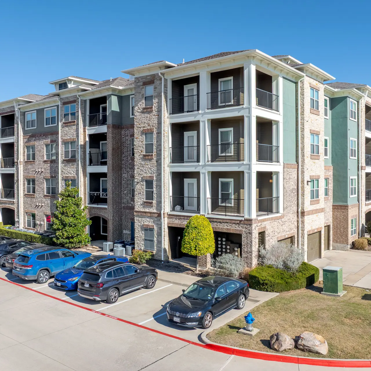 Building Exterior A modern apartment complex featuring multiple three to four-story buildings with a mix of stone and green exterior. A parking lot filled with cars is visible in front, along with some shrubs and small trees.