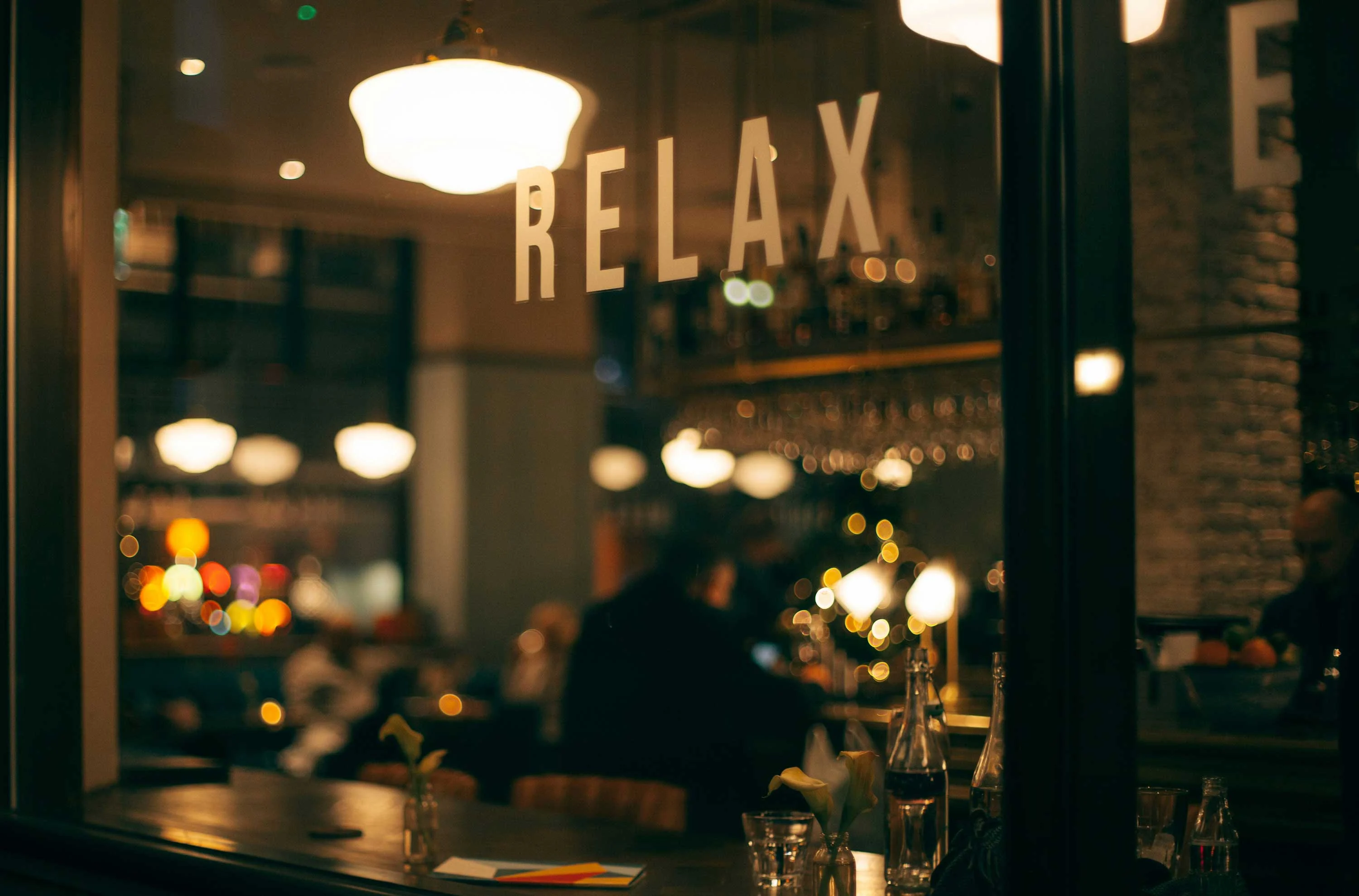 Interior of a bar or restaurant with a large window that has the word 'RELAX' displayed. Soft lighting and blurred figures of people can be seen inside, creating a warm and inviting atmosphere.
