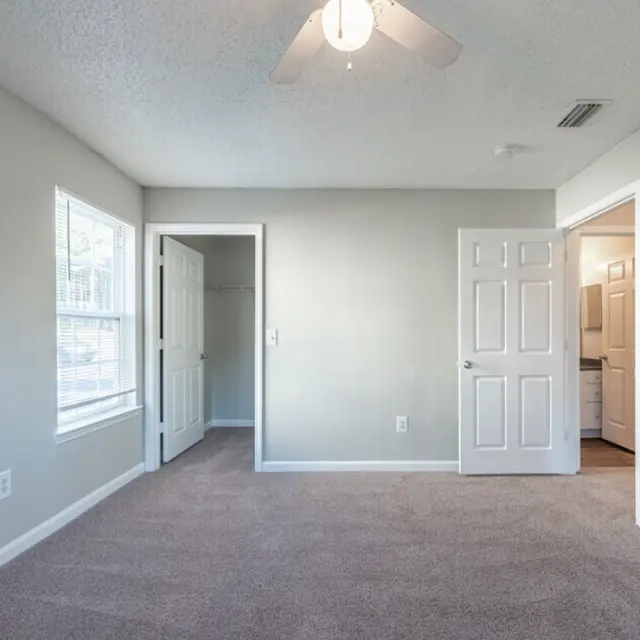 An empty room with carpeted flooring, white walls, and ceiling fan. It features two doors leading to other spaces and a window allowing natural light.