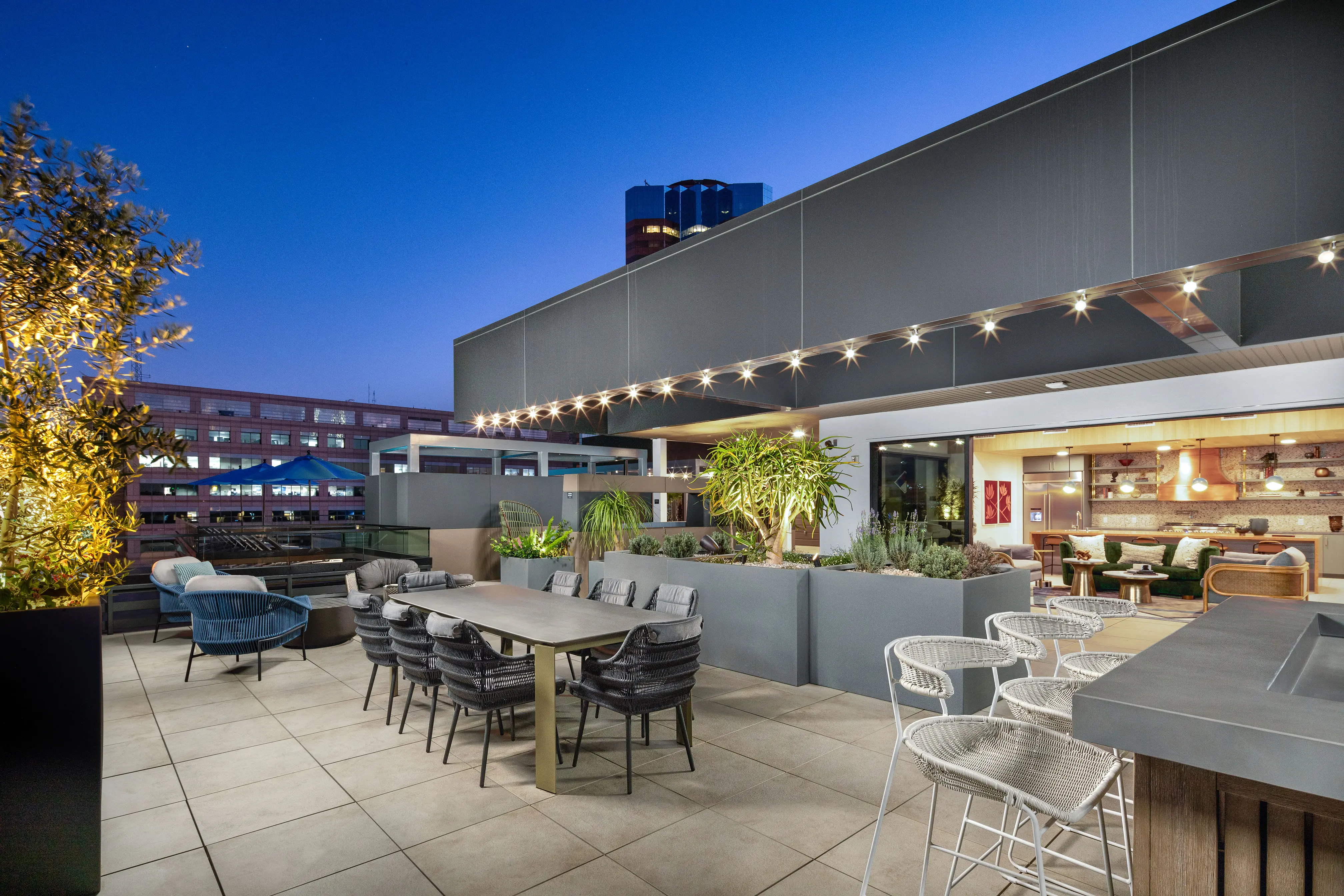 Modern Rooftop Space at Dusk A modern rooftop space with tables, chairs, and plants, illuminated by string lights at dusk.