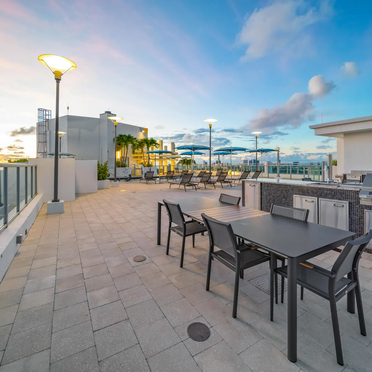 A spacious rooftop area featuring a modern dining setup with a table and chairs, barbecue grills, and a scenic backdrop of a colorful sunset.
