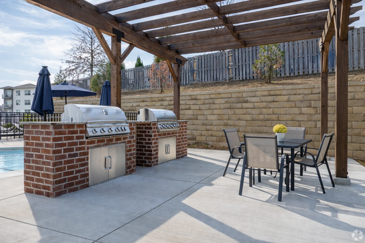 Outdoor grilling area featuring two stainless steel grills under a wooden pergola, accompanied by a dining table and poolside setting.