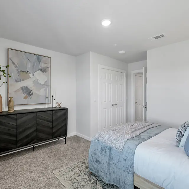 A modern bedroom featuring a bed with blue and white bedding, a black dresser with decorative vases, and a wall art piece. The room has light carpet and neutral-colored walls.