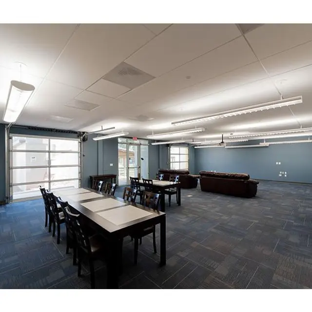 A spacious modern classroom featuring multiple tables and chairs, with a seating area that includes brown sofas. The room is well-lit with overhead lights and large windows allowing natural light to stream in.