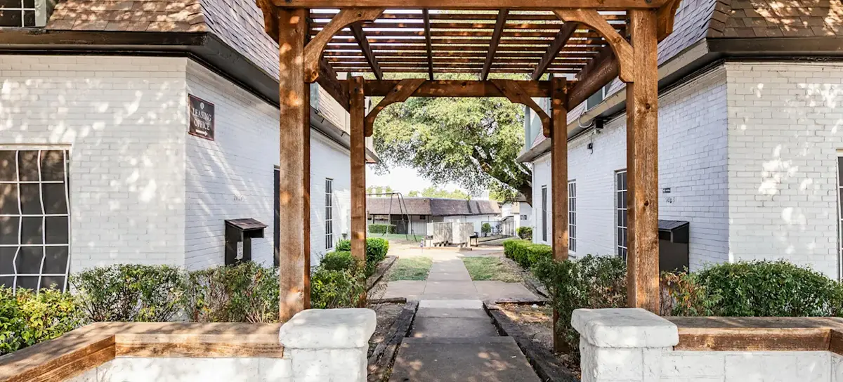 A wooden archway with a trellis leads to a pathway between white apartment buildings adorned with greenery.