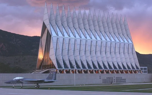 A modern architectural building with a series of pointed roofs resembling a cathedral against a sunset sky. In the foreground, a military jet is parked on the tarmac.