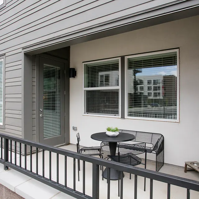 A modern patio with a round table, four chairs, and a potted plant, set against a gray exterior wall and large windows.