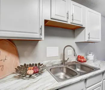 A modern kitchen countertop with white cabinets, a stainless steel sink, and decorative elements including a cutting board with text, a bowl holding small pumpkins, and a bowl of red apples.
