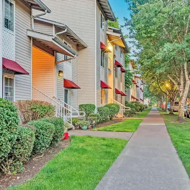 A well-maintained sidewalk lined with apartments featuring red awnings and neatly trimmed hedges. The scene includes a grassy area and parked cars along the roadside, with trees providing shade.