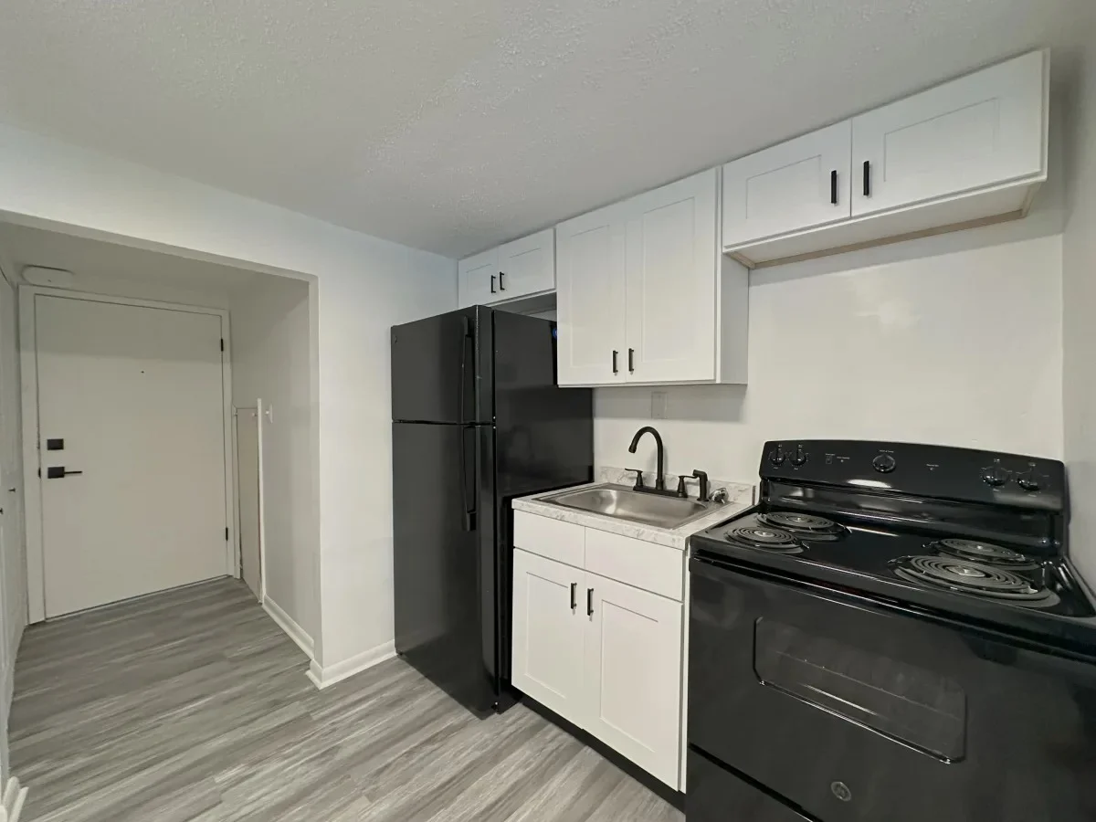 A modern kitchen with black and white cabinets, featuring a black refrigerator, stove, and sink. The kitchen has a light grey floor and is well-lit with an entrance visible in the background.