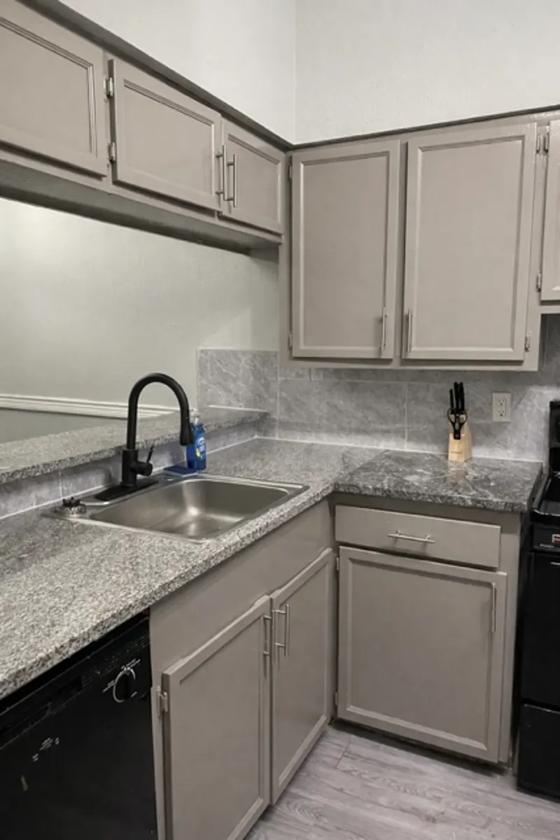A compact kitchen with light gray cabinets and dark countertops. The sink area includes a faucet and a small mirror above it. A black dishwasher is visible beneath the counter, and a stove is situated in the corner.