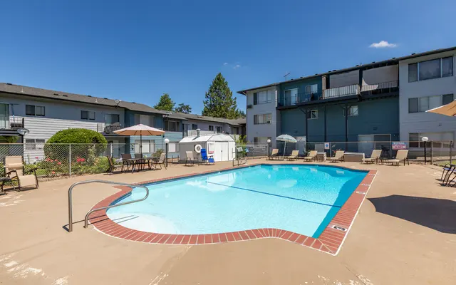 Apartment Complex Pool Area A swimming pool surrounded by lounge chairs, umbrellas, and an apartment building in the background on a sunny day.