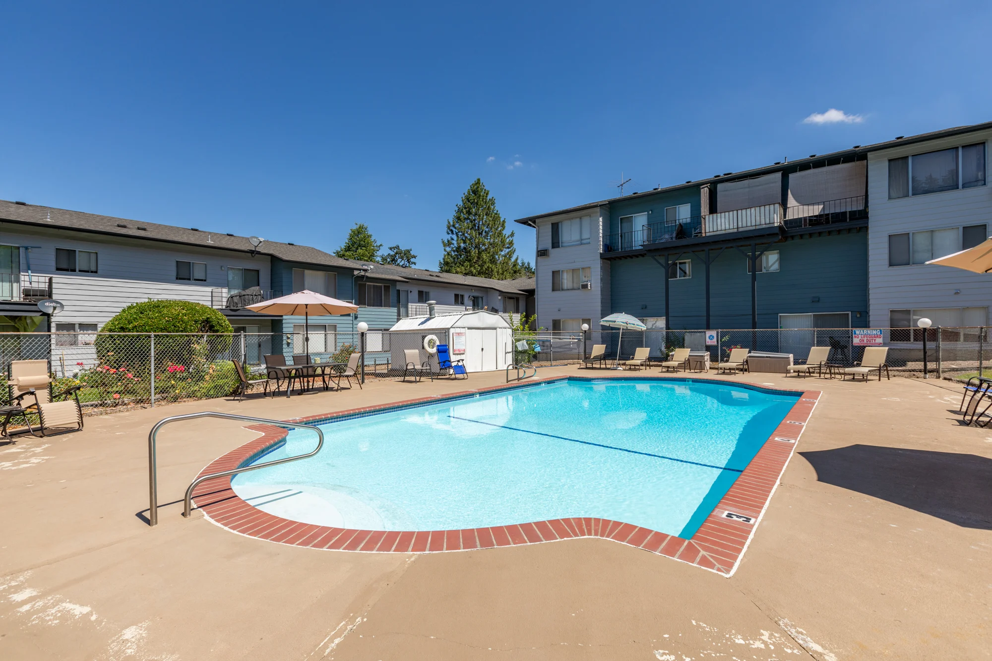 Apartment Complex Pool Area A swimming pool surrounded by lounge chairs, umbrellas, and an apartment building in the background on a sunny day.