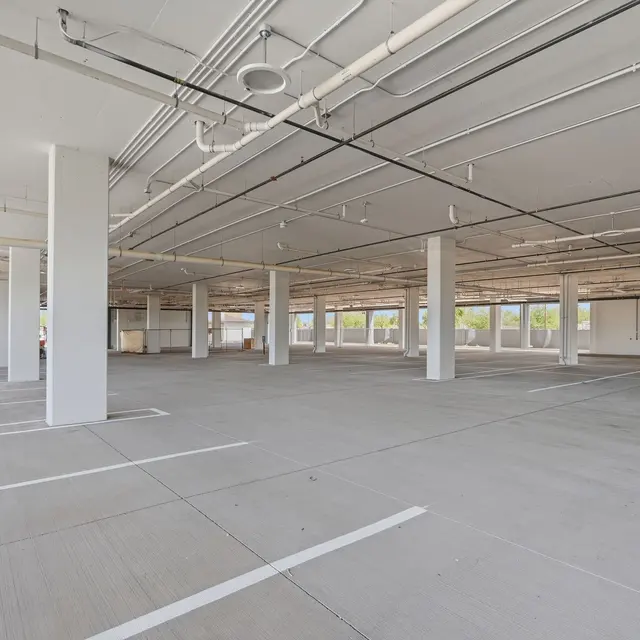 Empty Parking Garage Interior An empty parking garage interior with concrete flooring and white pillars, featuring a few parked cars and large windows on one side.