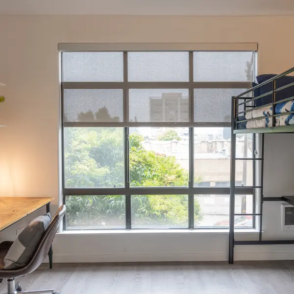 A modern bedroom featuring a bunk bed, a study desk, and large windows allowing natural light. A small potted plant is on the desk, and greenery is visible outside.