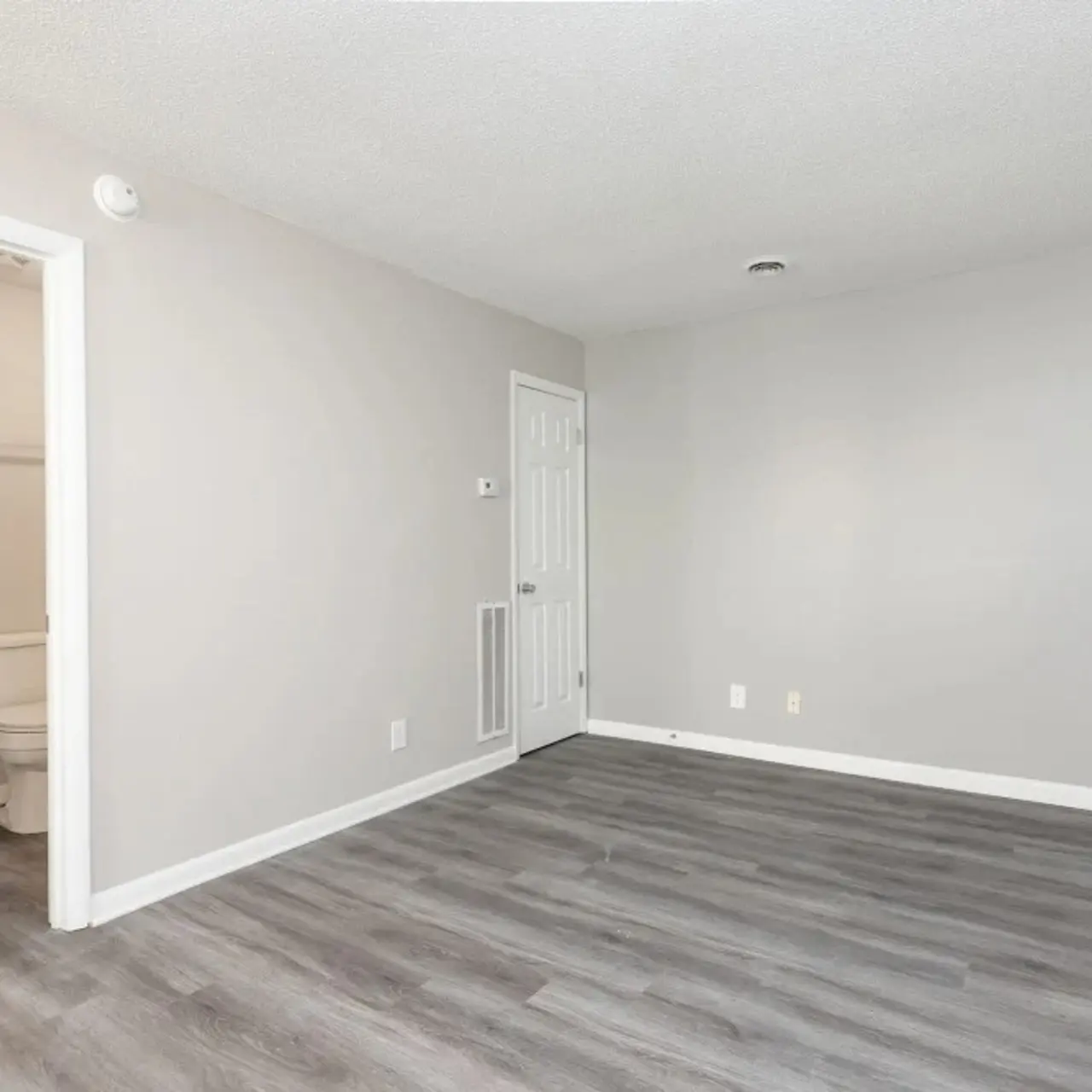 A spacious empty room with gray walls and laminate flooring, showcasing natural light. On the left, there is an open door leading to a bathroom with a sink and toilet visible. The room has no furniture, making it feel open and airy.