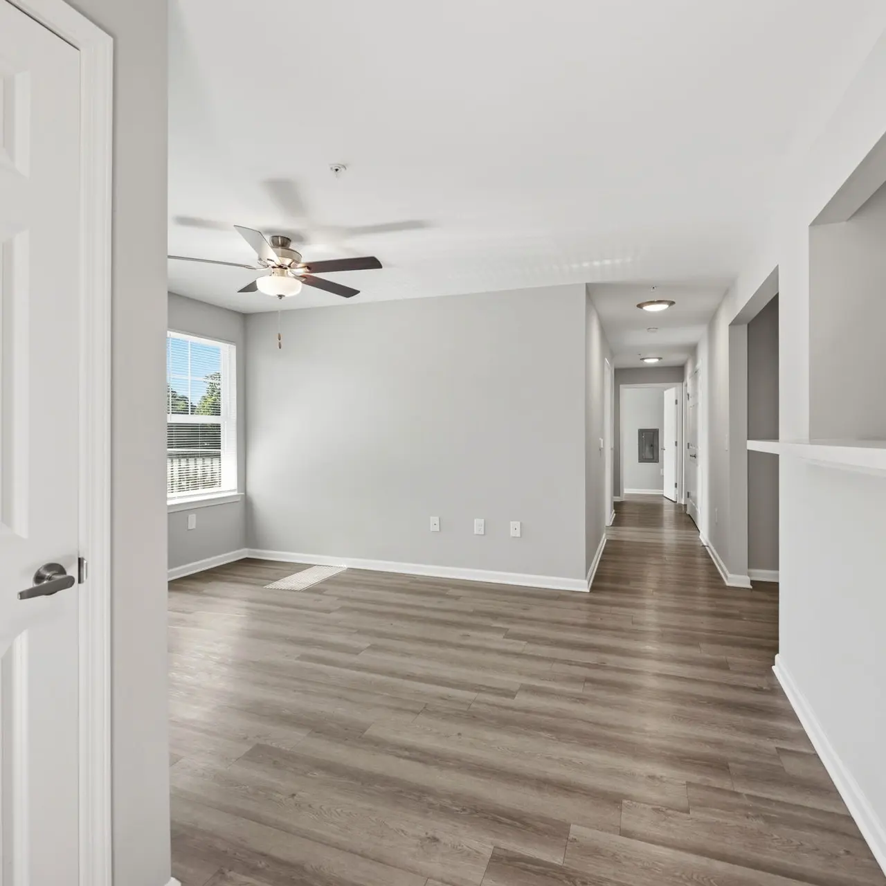 A modern interior view of a living space featuring light grey walls, hardwood flooring, and a ceiling fan. There are windows allowing natural light, and a layout showing an open area leading into another room.