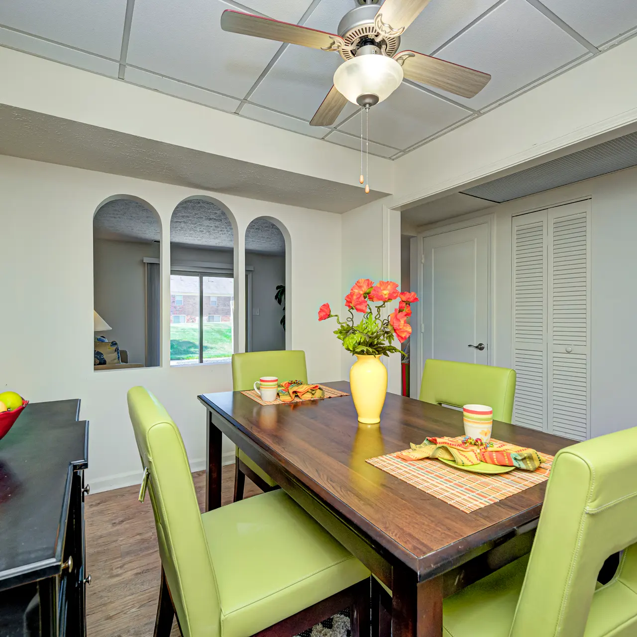 Cozy Dining Area A dining area featuring a wooden table surrounded by green chairs, with a vase of flowers on the table and a fruit bowl in the background. There are large windows allowing natural light into the room.
