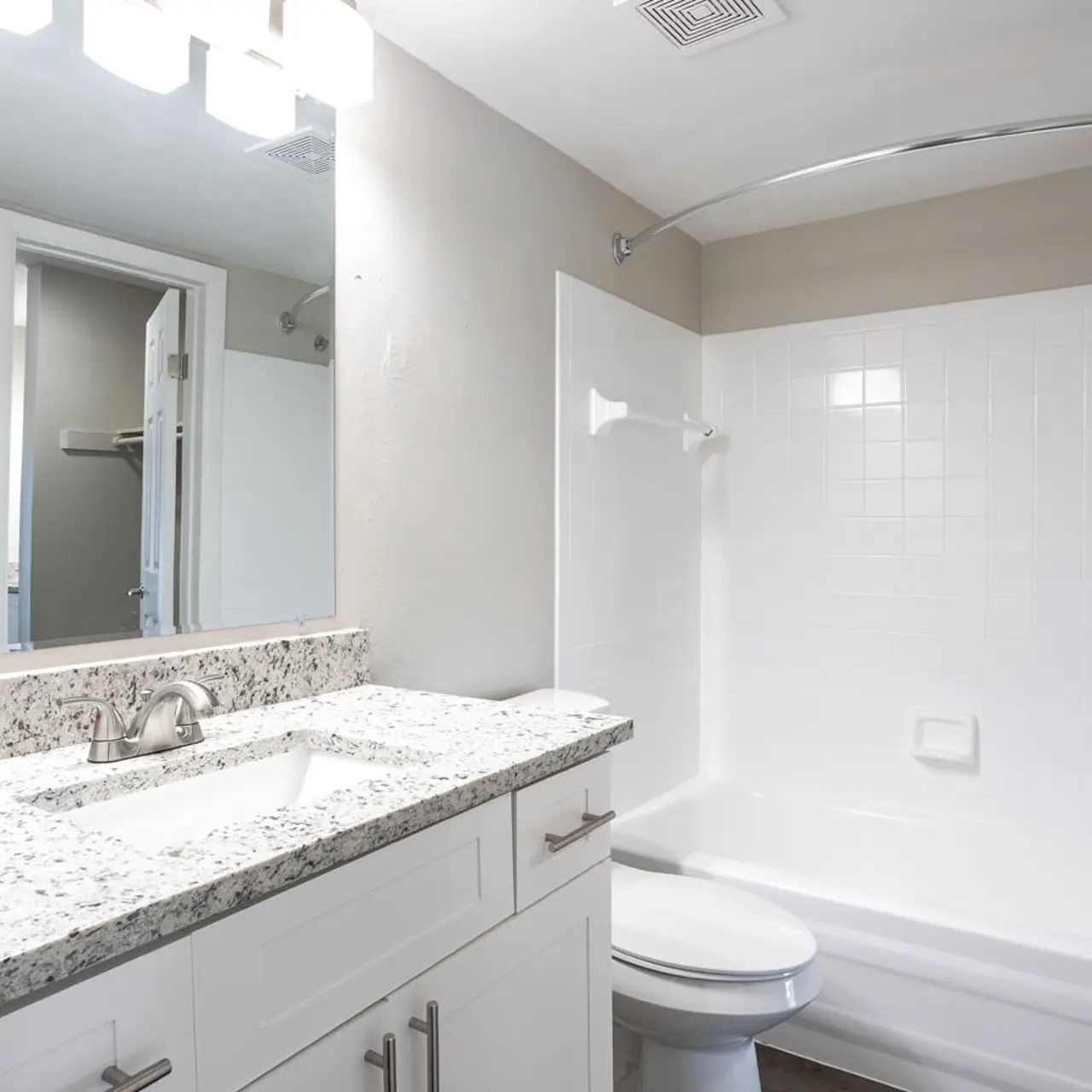 A modern bathroom featuring a granite countertop with a sink, a mirror above, and a bathtub with a shower curtain. The walls are painted in a light gray color and there is a door leading to another room visible in the background.