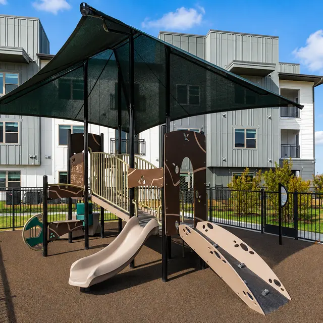 A playground with a slide and climbing structure under a shade canopy, surrounded by a fenced area and modern apartment buildings in the background.