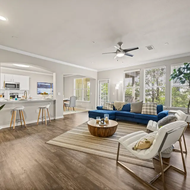 Contemporary Living Room A modern living room featuring a blue sofa, two white chairs, and a wooden coffee table on a beige rug. The room has large windows allowing natural light and greenery visible outside, with light wood flooring and a ceiling fan.