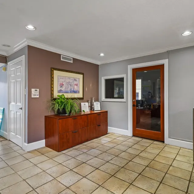 A bright and spacious home entryway featuring tiled flooring, a wooden sideboard with plants, and a decorative wall art. A doorway in the background leads to a pool area and another room is visible through a glass door.