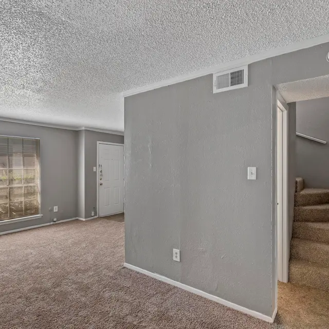 A spacious living room with light grey walls and beige carpet, featuring a large window with sheer curtains and stairs leading to an upper level.