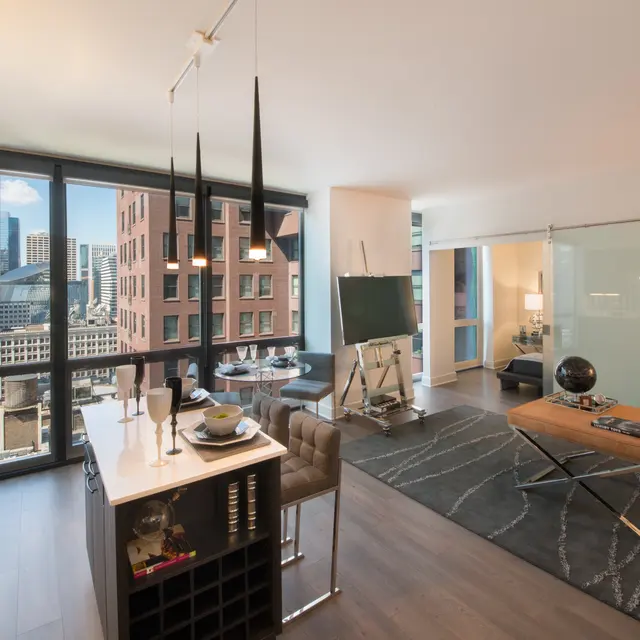 A modern loft apartment featuring large windows with city views, a stylish dining area, and contemporary furniture. The space includes a kitchen island with seating, a wooden coffee table, and a rug on the hardwood floor.