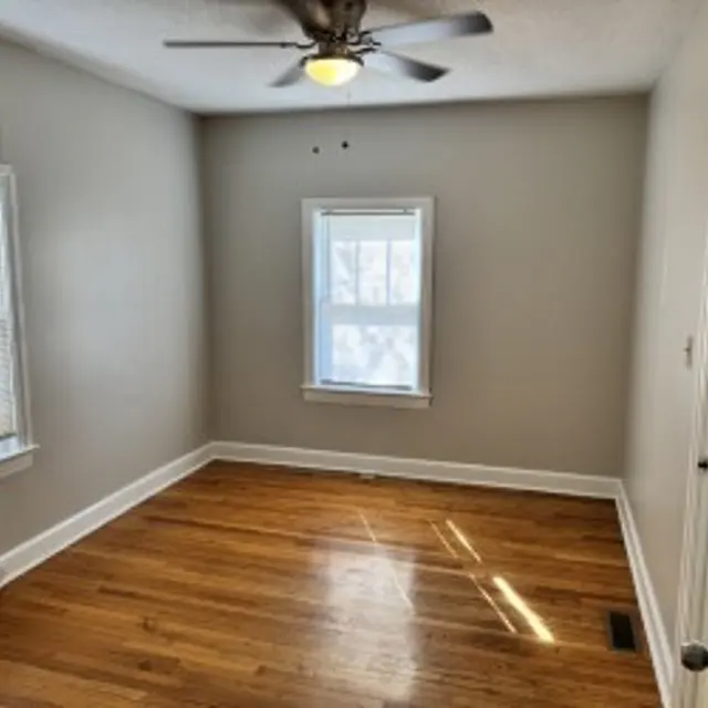 A small, empty room with light gray walls and hardwood flooring. It features one window with blinds, a small window, and a ceiling fan.