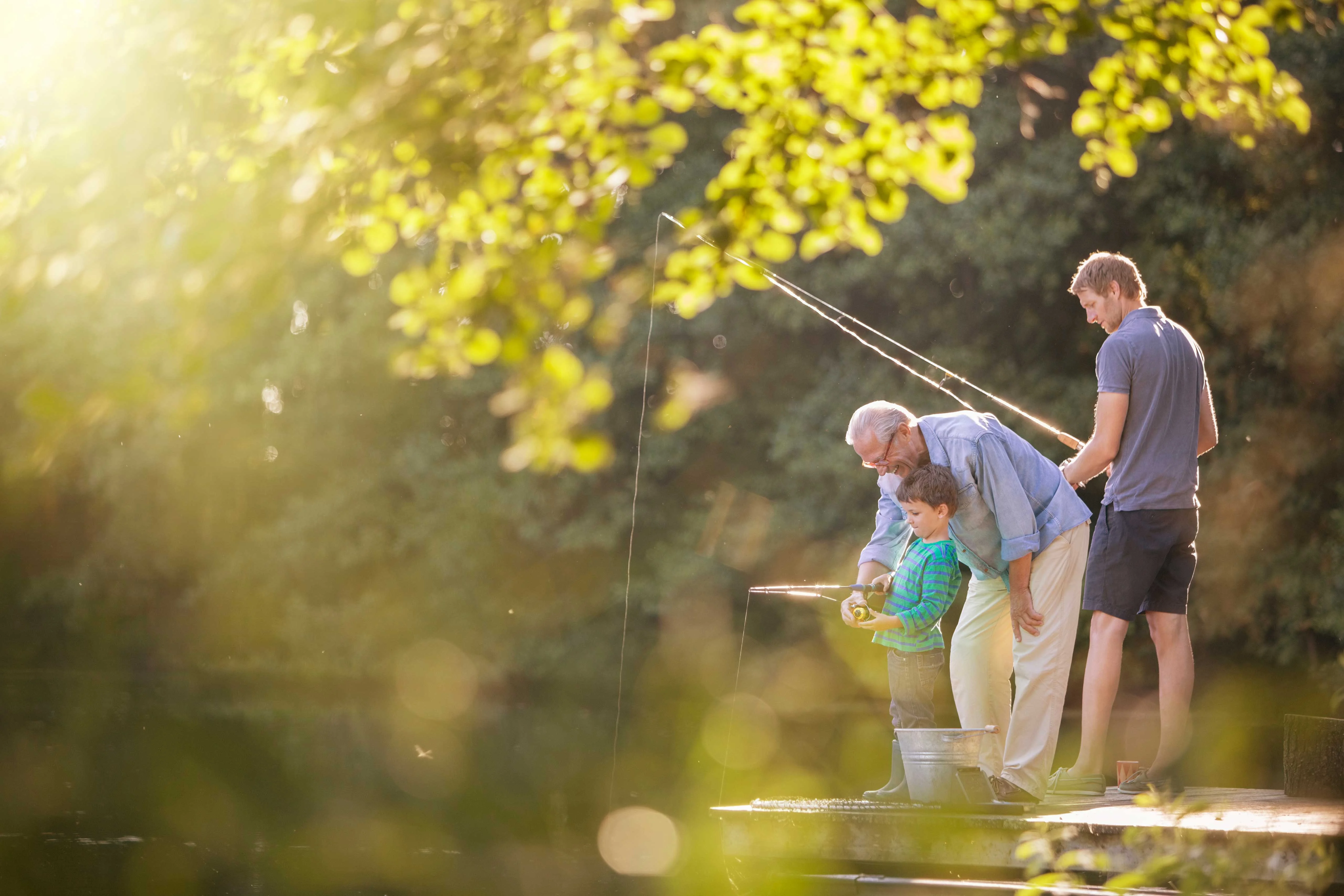A grandfather, father, and young boy fishing together on a sunny day by the water. The scene is illuminated by soft sunlight filtering through the trees, creating a warm atmosphere.