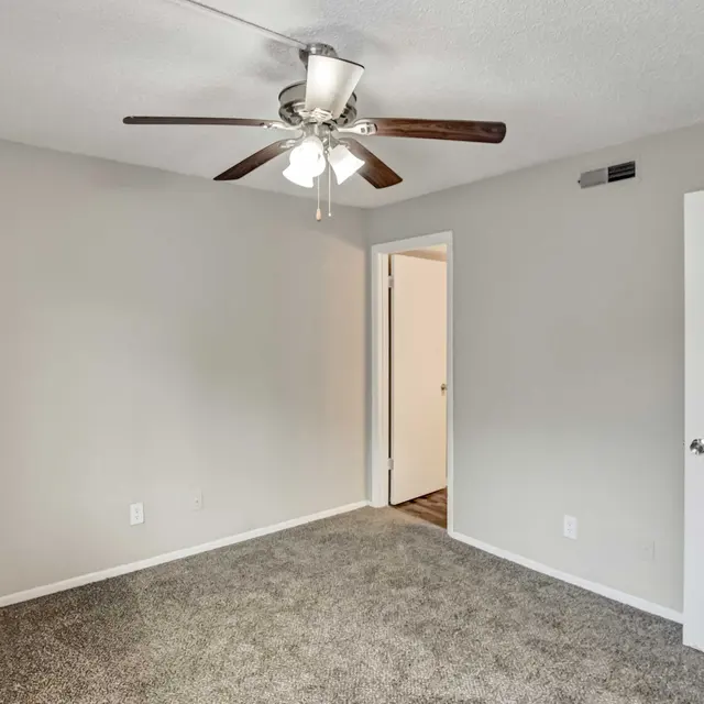 A sparsely furnished room with gray walls and carpet, featuring a ceiling fan and a door leading to another room.