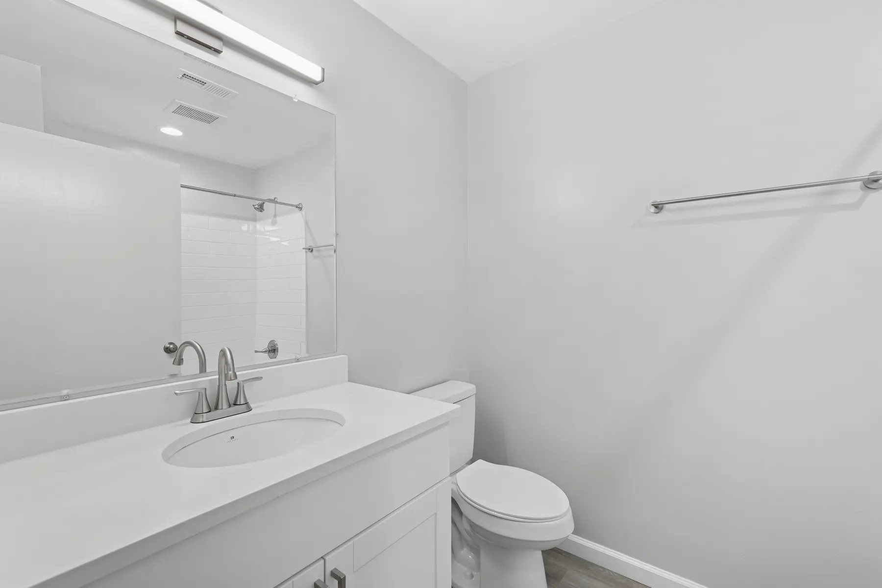 A modern bathroom featuring a white countertop with a sink, a toilet, and a walk-in shower. The walls are light gray, and the space is well-lit with a mirror above the sink.