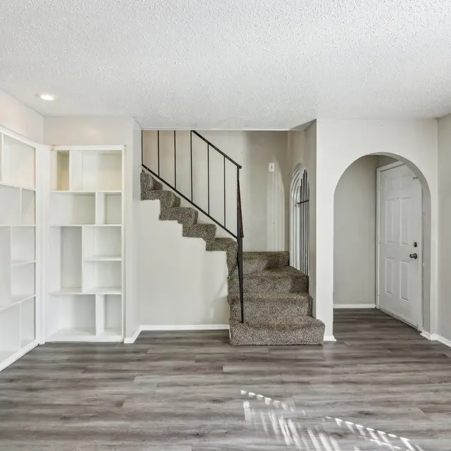 A spacious living room area featuring light-colored walls, wooden flooring, and carpeted stairs leading to an upper level. A white shelving unit is built into one wall, with a small entrance visible on the right.