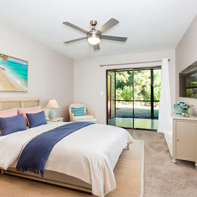 A modern bedroom with a ceiling fan, featuring a king-size bed with light bedding and decorative pillows. There are two lamps on nightstands beside the bed, an armchair, and large glass doors leading to an outdoor view.