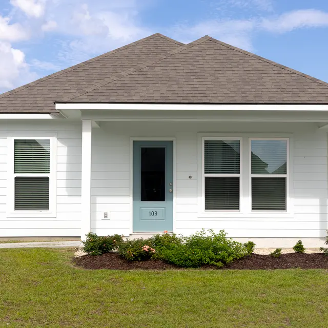 Front View of a Modern House A single-story white house with a gray roof and blue front door. The yard is well-kept with some plants and flowers, and a wooden fence is visible on one side.