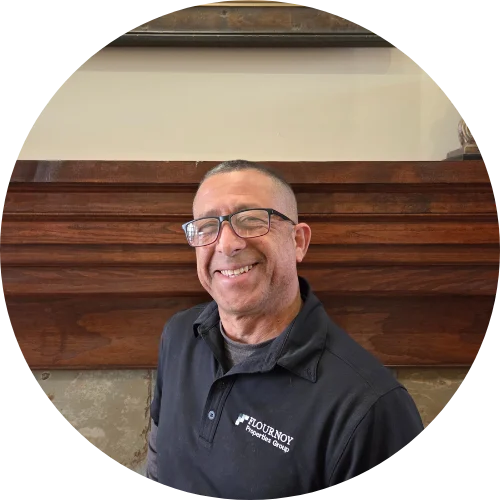 A smiling man wearing glasses and a dark polo shirt stands in front of a wooden mantle.