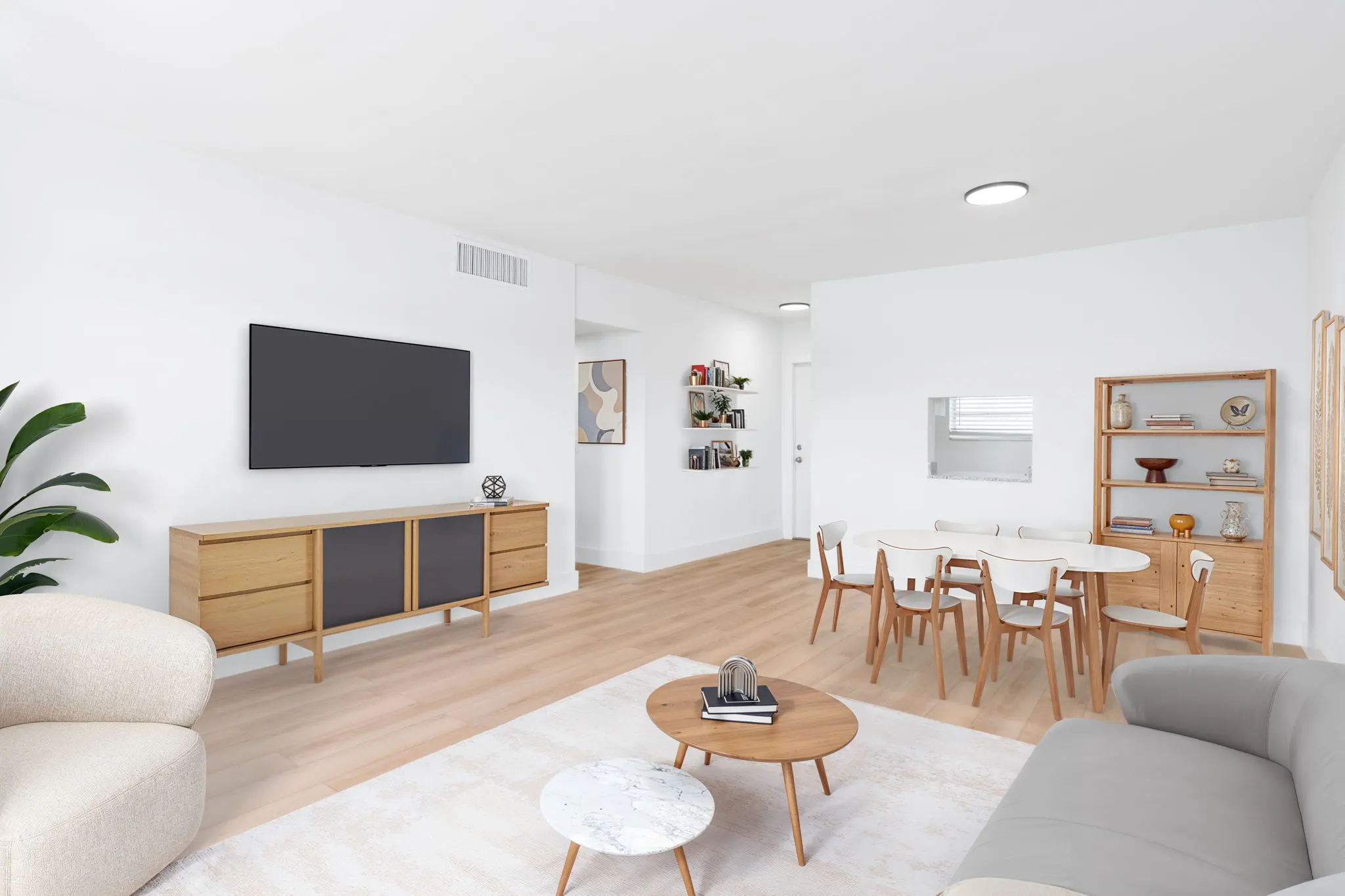 A modern living room featuring light wood floors and white walls, with a beige sofa, a gray couch, a round marble coffee table, and a wooden dining table with chairs. A flat-screen TV is mounted on the wall, and decorative items are displayed on a nearby shelf.