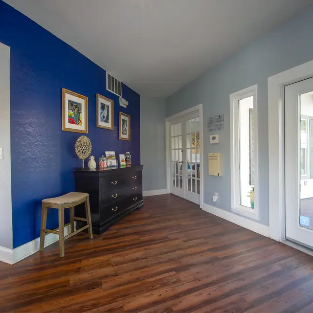 A spacious hallway featuring a blue accent wall with framed pictures, a dark wooden dresser, a stool, and a glass door leading outside.