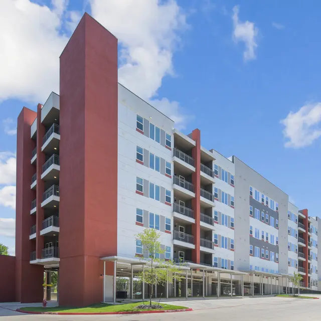 A modern apartment building with a mix of red and white exterior, featuring several balconies and large windows under a bright blue sky with a few clouds.