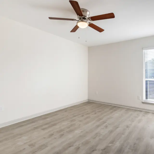 An empty room with a ceiling fan and large window, featuring light wood flooring and white walls.