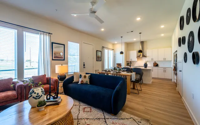 A modern apartment living room featuring a blue sofa, two red armchairs, a wooden table, and a kitchen area in the background.