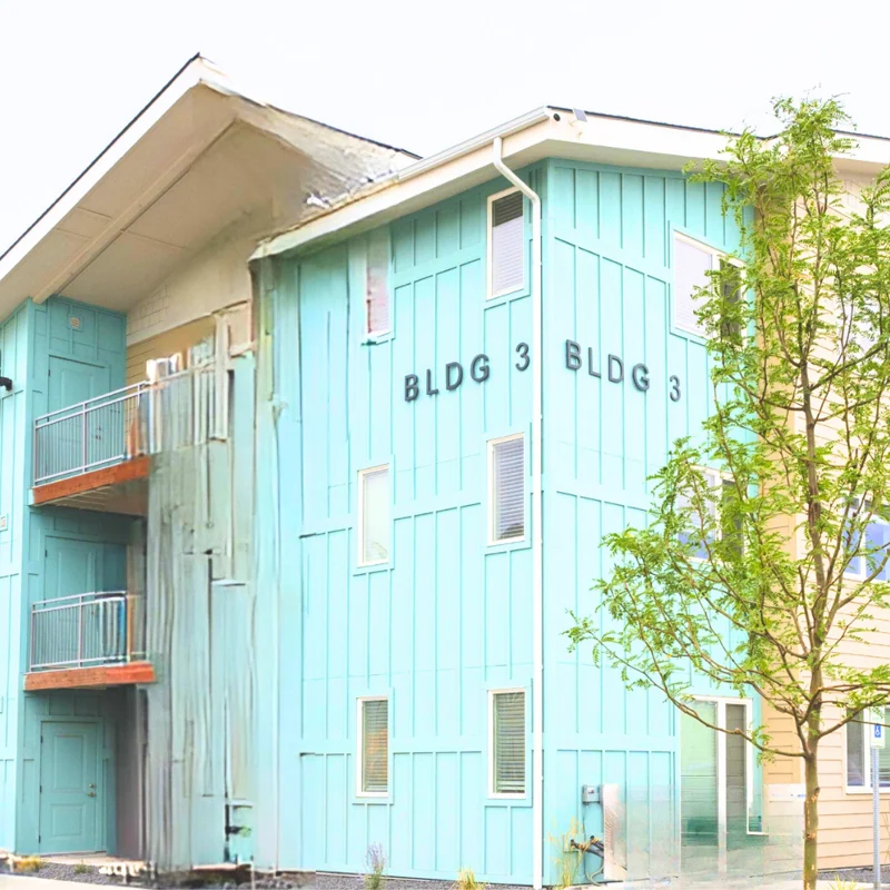 Residential Building BLDG 3 A multi-story residential building with a light blue exterior, labeled 'BLDG 3'. The building features multiple balconies and large windows, surrounded by trees and greenery.