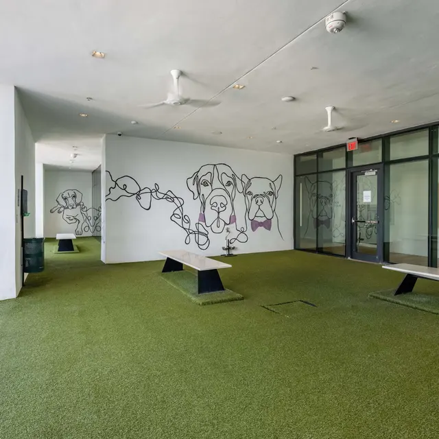 An indoor dog park area featuring artificial turf, large glass windows, and a mural of dogs on the wall, complemented by minimal seating.