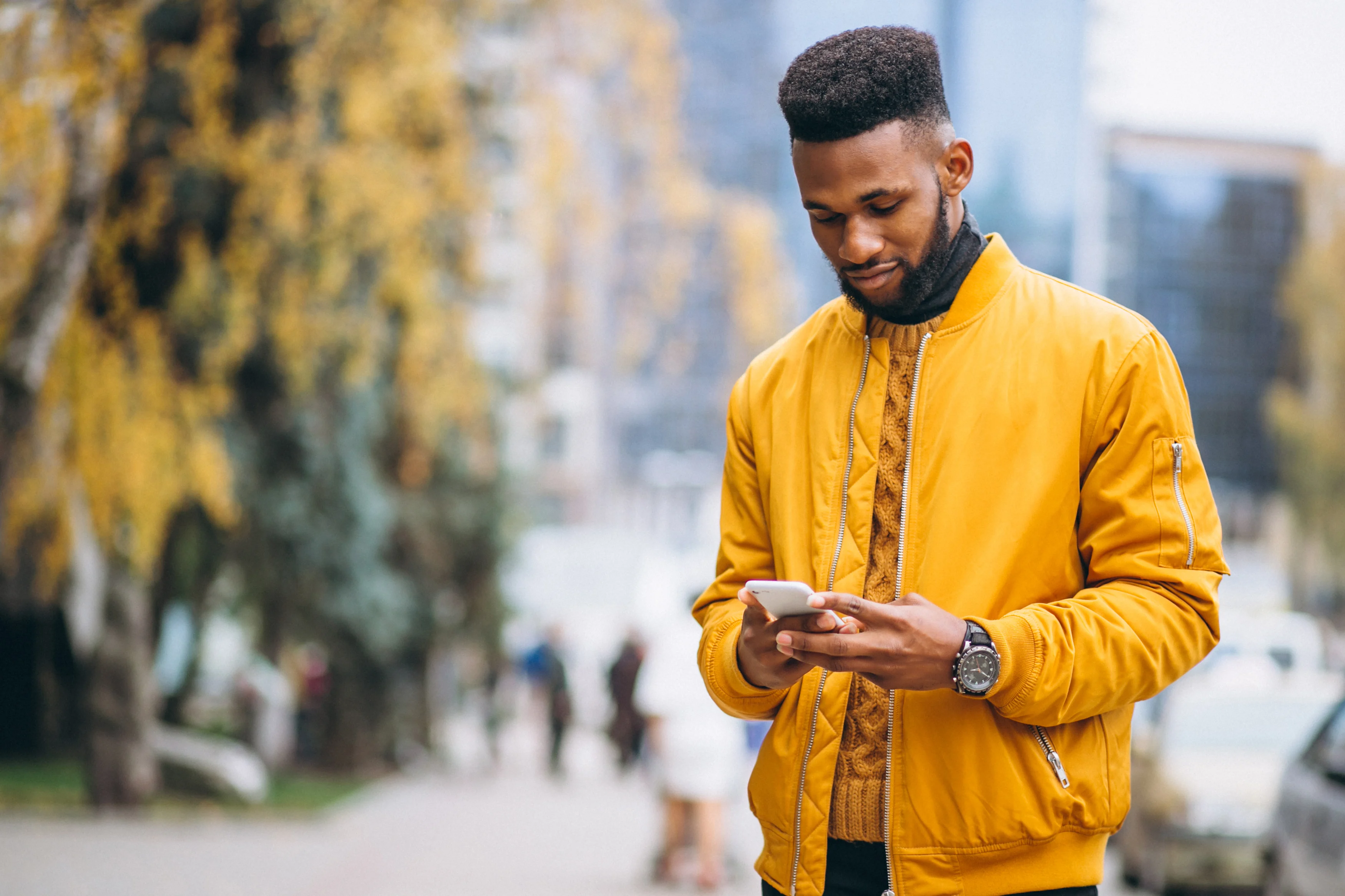 A man wearing a yellow jacket stands outdoors, looking at his smartphone with a thoughtful expression. The background features autumn foliage and blurred figures walking in a city setting.