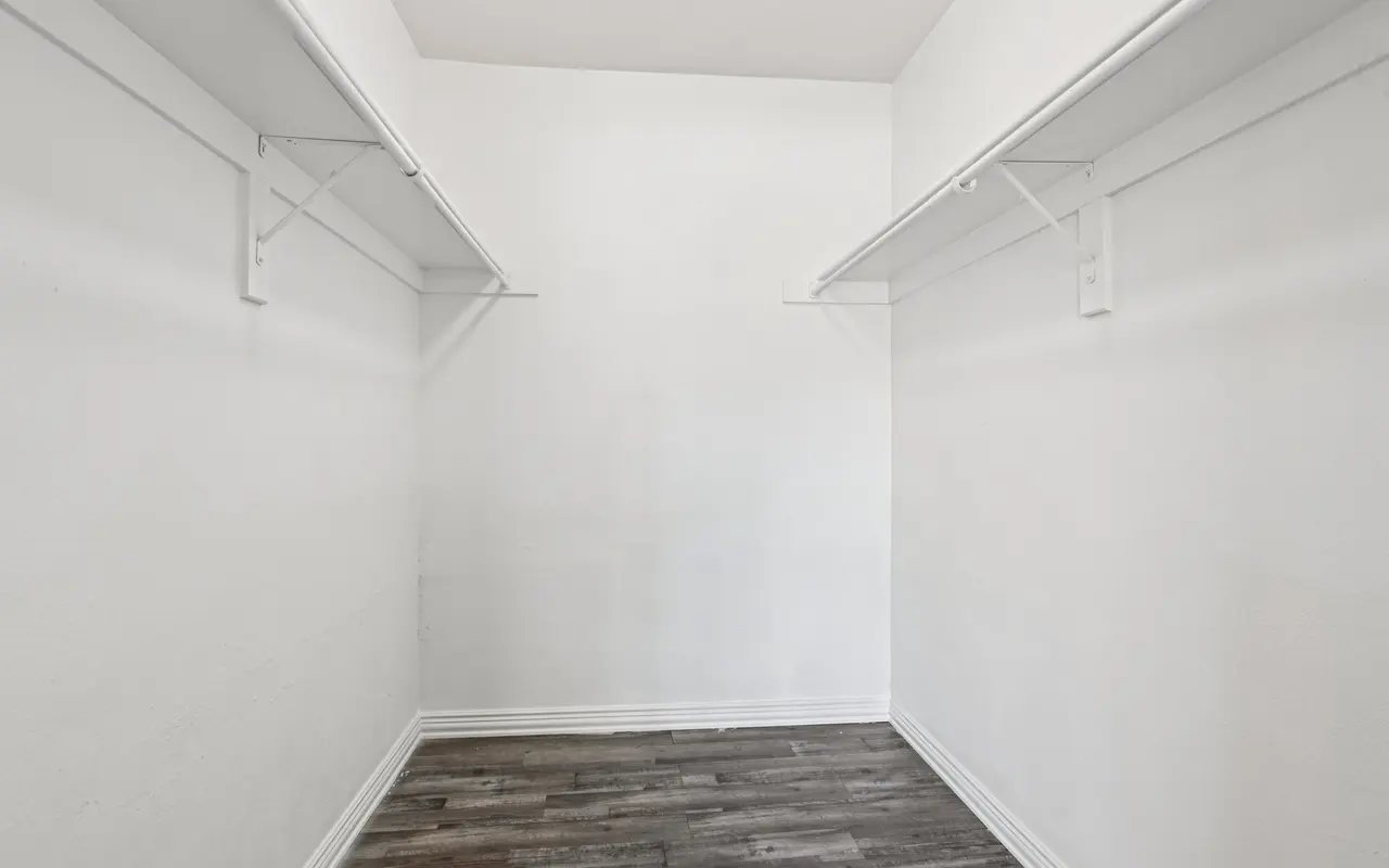 An empty closet with white walls and wooden flooring, featuring two shelves on opposite sides.