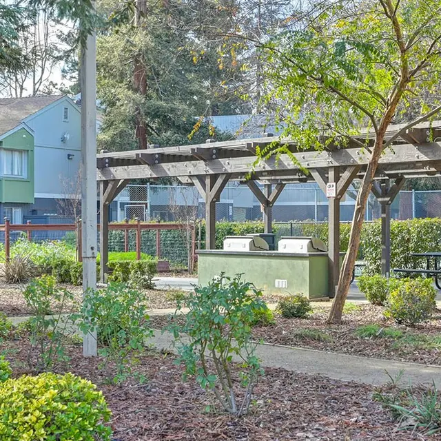 A landscaped park area featuring a wooden pavilion with picnic tables, surrounded by green shrubs and bushes.