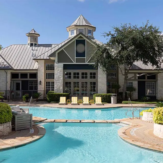 A resort-style swimming pool surrounded by a landscaped area with palm trees and colorful plants, leading up to a large building with a distinctive architectural style, featuring a peaked roof and many windows.