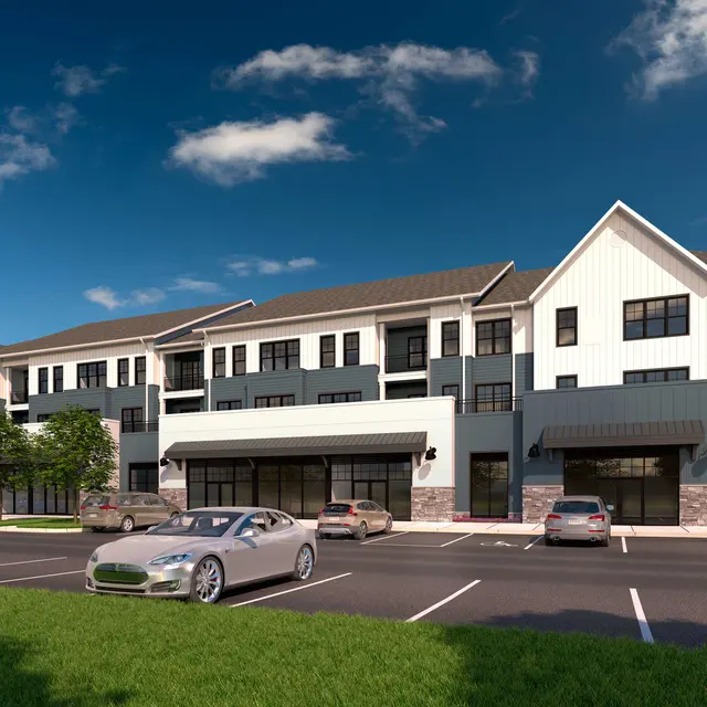 A modern multi-story building with a mix of white and gray siding, featuring balconies and large windows. Several cars are parked in front of the building, and there are trees and grassy areas nearby, under a blue sky with some clouds.