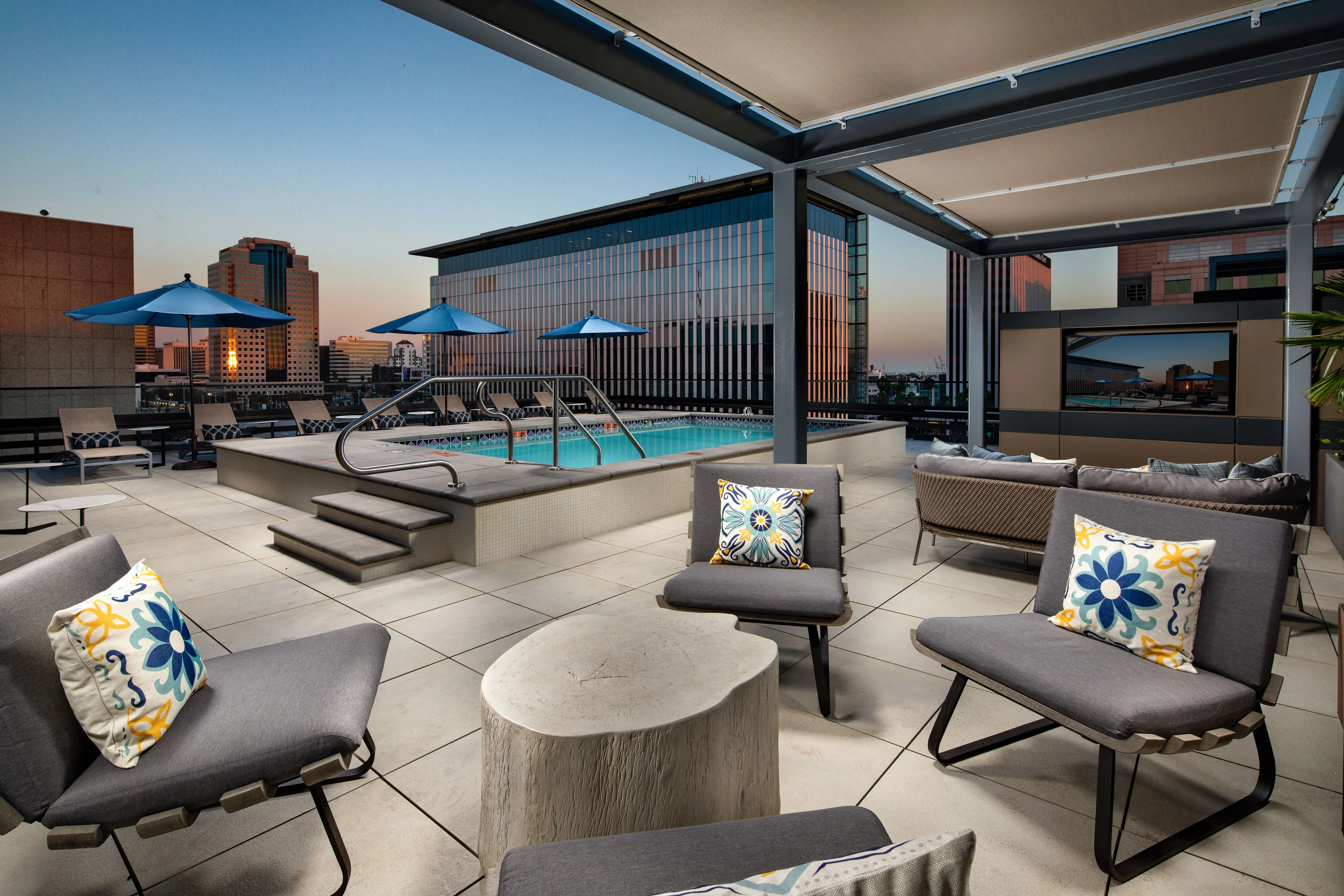 A modern rooftop pool area featuring comfortable lounge seating, a swimming pool, and city views at sunset.